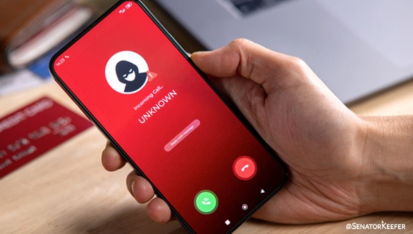 Hand holding a smartphone displaying an incoming call screen with a red background, showing an unknown caller represented by a masked icon. A laptop and a credit card sit on the table in the background.