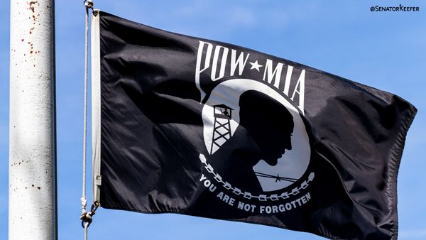 Black POW/MIA flag flying against a blue sky, featuring the silhouette of a guarded figure inside a white circle, with the words 'POW/MIA' at the top and 'You Are Not Forgotten' at the bottom.
