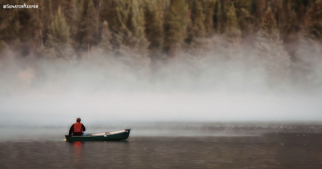 A person wearing a red vest in a canoe on a lake surrounded by mist.