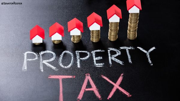 stacks of coins arranged in increasing height across a dark surface, each topped with a small red-and-white house figurine. Below the stacks, the words “PROPERTY TAX” are written in chalk.